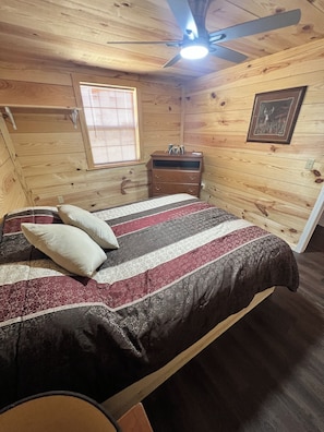 A wooden-paneled room with a bed, a ceiling fan, a window with blinds, a dresser, and a framed picture on the wall.