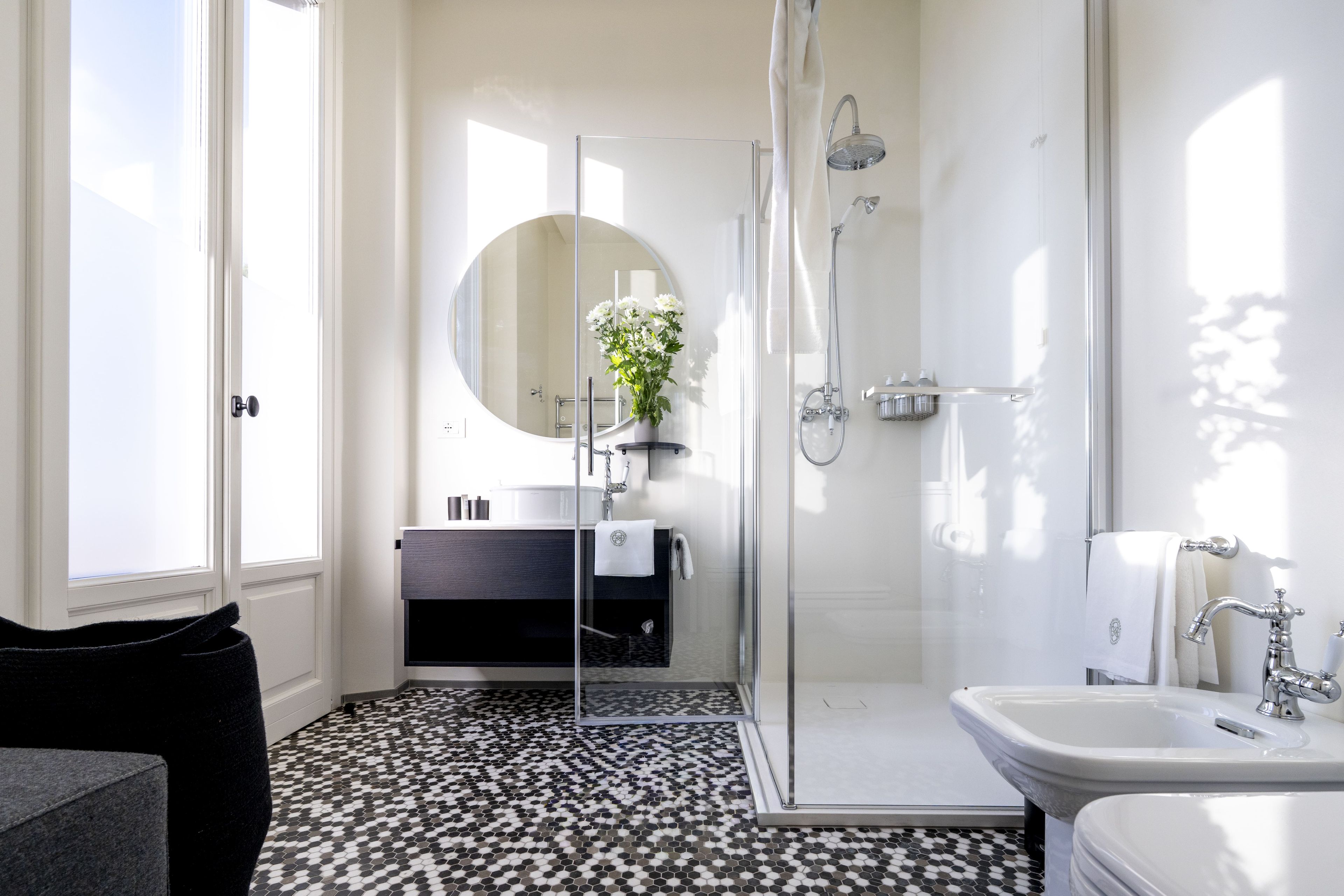 A modern bathroom with a glass shower enclosure, a round mirror, and a black and white patterned floor.