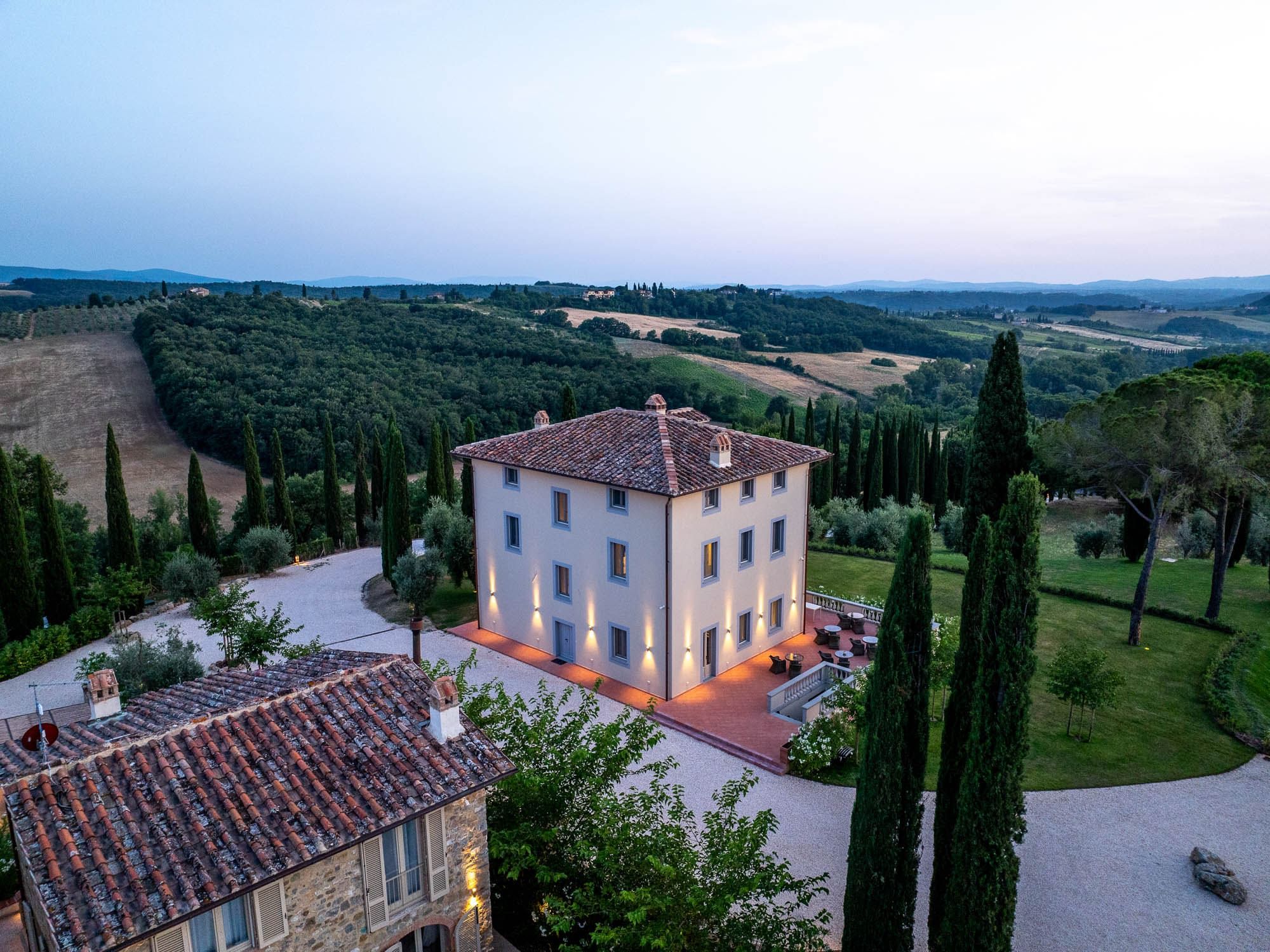 A two-story building with a tiled roof, surrounded by a well-maintained garden and tall cypress trees.