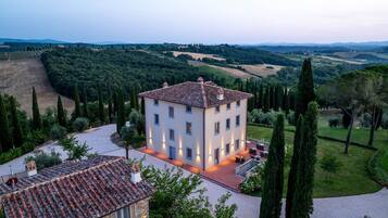 A two-story building with a tiled roof, surrounded by a well-maintained garden and tall cypress trees.