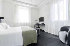 Ett hotellrum med en säng, ett skrivbord, en stol, en tv och stora fönster med tunna, genomskinliga gardiner.