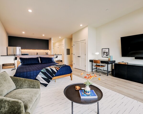 A modern hotel room with a bed, a desk, a TV, and a coffee table with a book and flowers.
