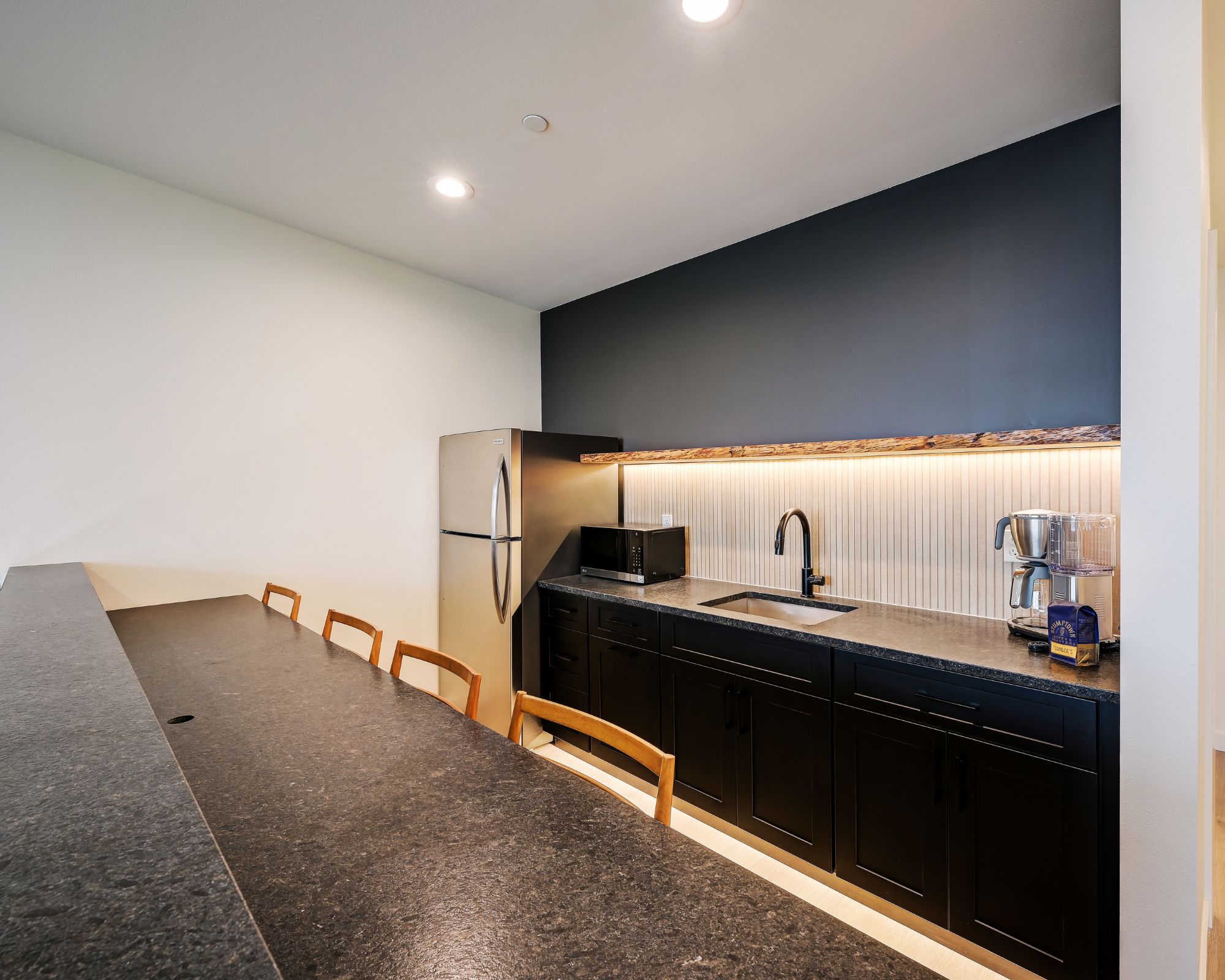 Une cuisine moderne avec un revêtement mural foncé, un réfrigérateur en acier inoxydable, un four à micro-ondes, un évier avec un robinet et une machine à café.