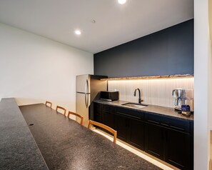 A modern kitchen with a dark backsplash, a stainless steel refrigerator, a microwave, a sink with a faucet, and a coffee maker.