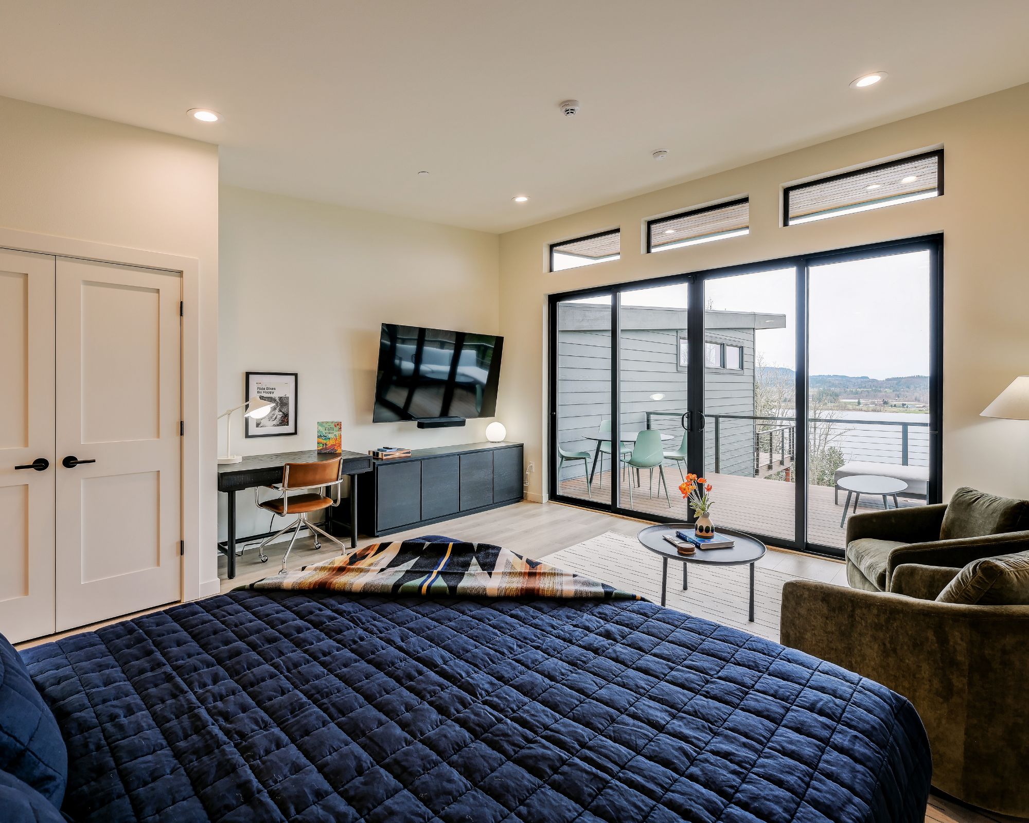 A modern bedroom with a large bed, a desk, a TV, and a balcony with a view.