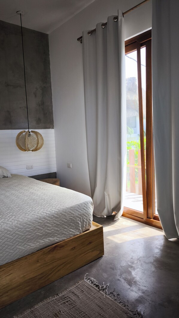 Un dormitorio con cama, lámpara colgante, ventana con cortinas y vista a follaje exterior.