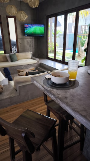 A modern living room with a sofa, a dining area with a table and chairs, a television, and a view of the outdoors.