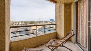 Varanda com vista para um parque de estacionamento e edifícios, uma cadeira de madeira e uma janela com cortina estampada.