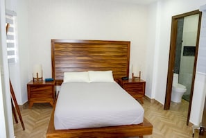 A neatly made bed with a wooden headboard, two bedside tables with lamps, and a bathroom visible through an open door. - Allende Heights: 1BR Oasis w/ Rooftop Pool (Puerto Vallarta)