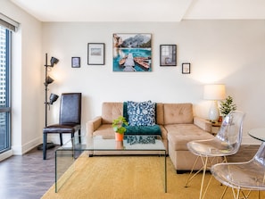 A modern living room with a glass coffee table, a beige sofa, a clear glass table, and a chair. There are framed pictures on the wall, a floor lamp, and a window with blinds. - StayToronto - The Britt Suites (Toronto)