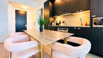 A modern kitchen with dark cabinets, a marble countertop, and a dining area with a wooden table and white upholstered chairs.