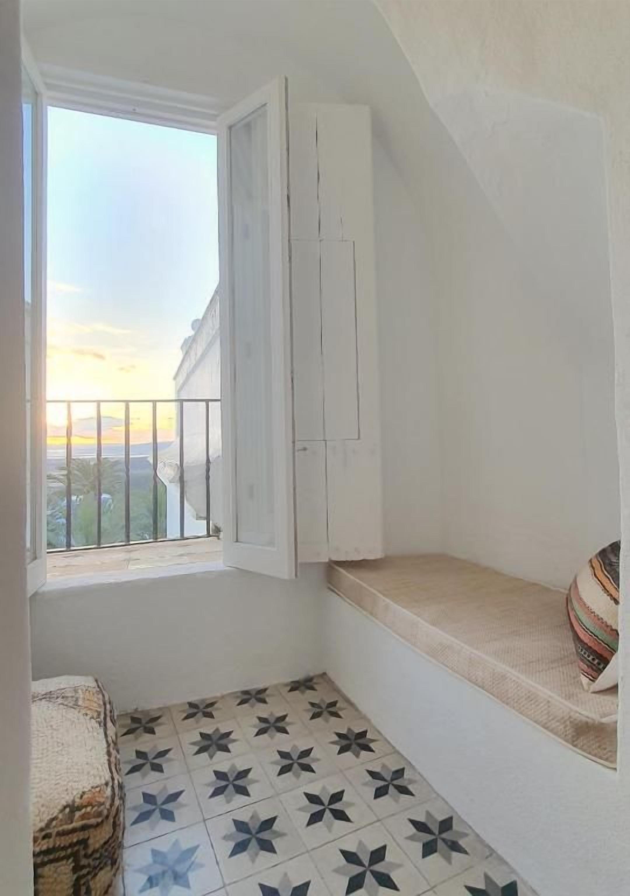 Un balcón pequeño con barandilla blanca, una ventana con cortinas, suelo de baldosas y un banco.