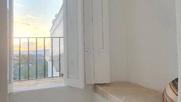 Un balcón pequeño con barandilla blanca, una ventana con cortinas, suelo de baldosas y un banco.