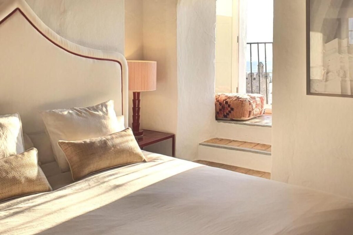 A bedroom with a bed, pillows, a nightstand, a lamp, a window with a view, and a framed picture on the wall.