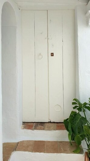 A white door with a small brass plate, a potted plant to the right, and a tiled floor. - Canalejas 3 Vejer (Vejer de la Frontera)