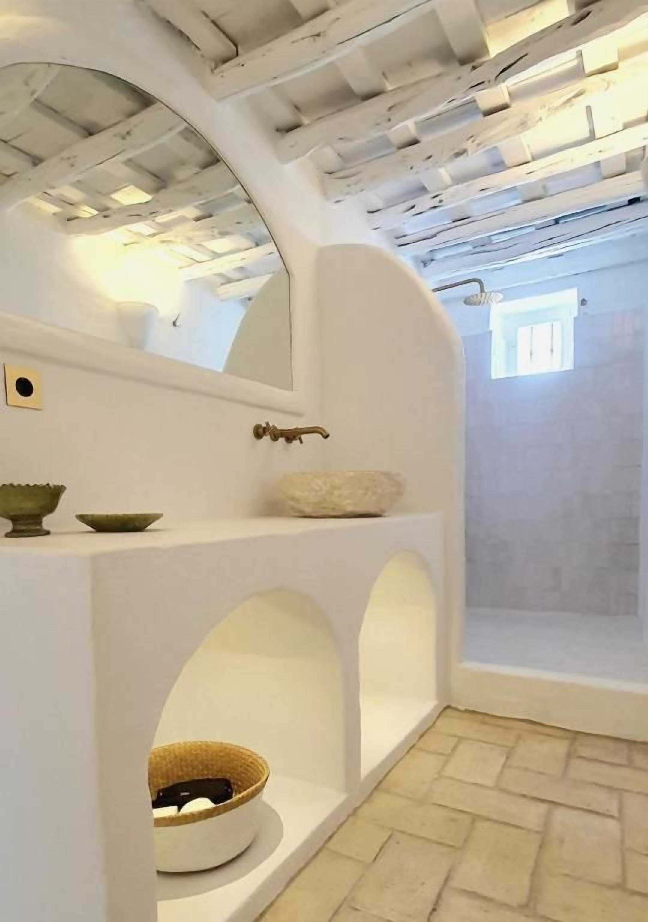 Una habitación con paredes blancas, suelo de piedra, un ventanal pequeño y una estantería con cuencos.