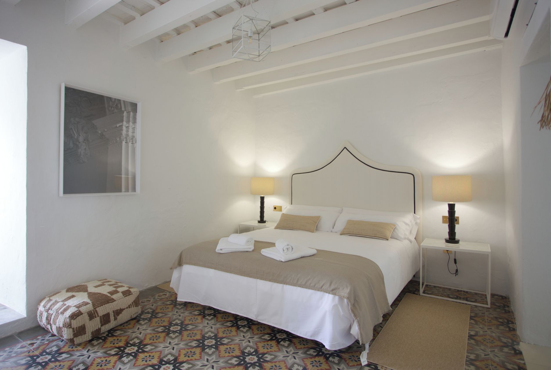 A bedroom with a bed, two bedside tables, a nightstand, a patterned rug, and a framed picture on the wall.