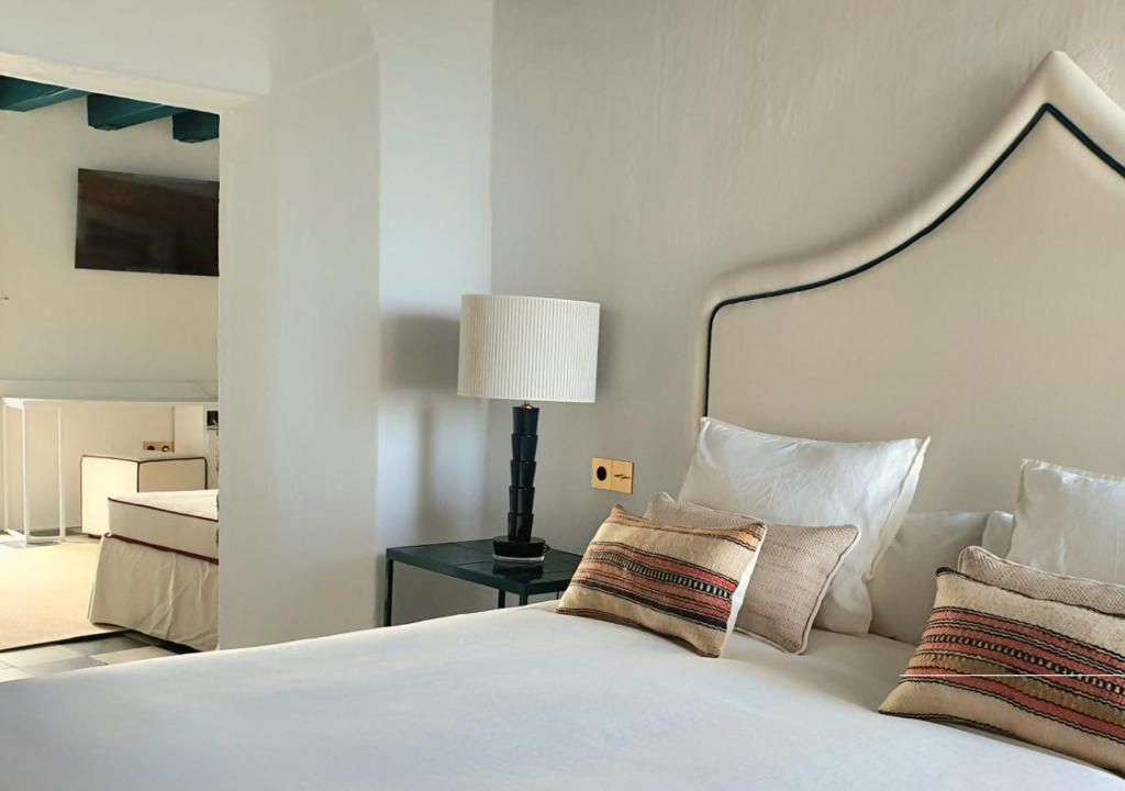 Habitación de hotel con cama, mesita de noche con lámpara, espejo y televisor montado en la pared.