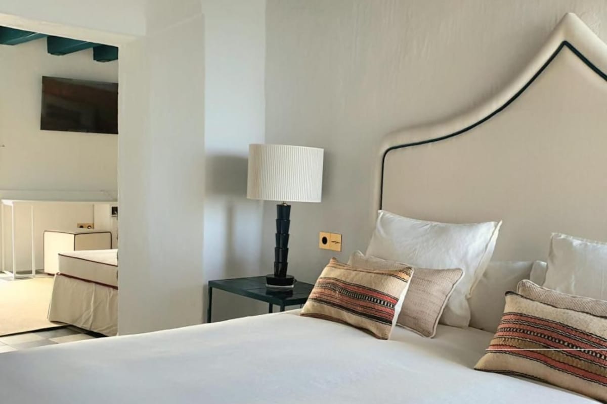 Habitación de hotel con cama, mesita de noche con lámpara, espejo y televisor montado en la pared.