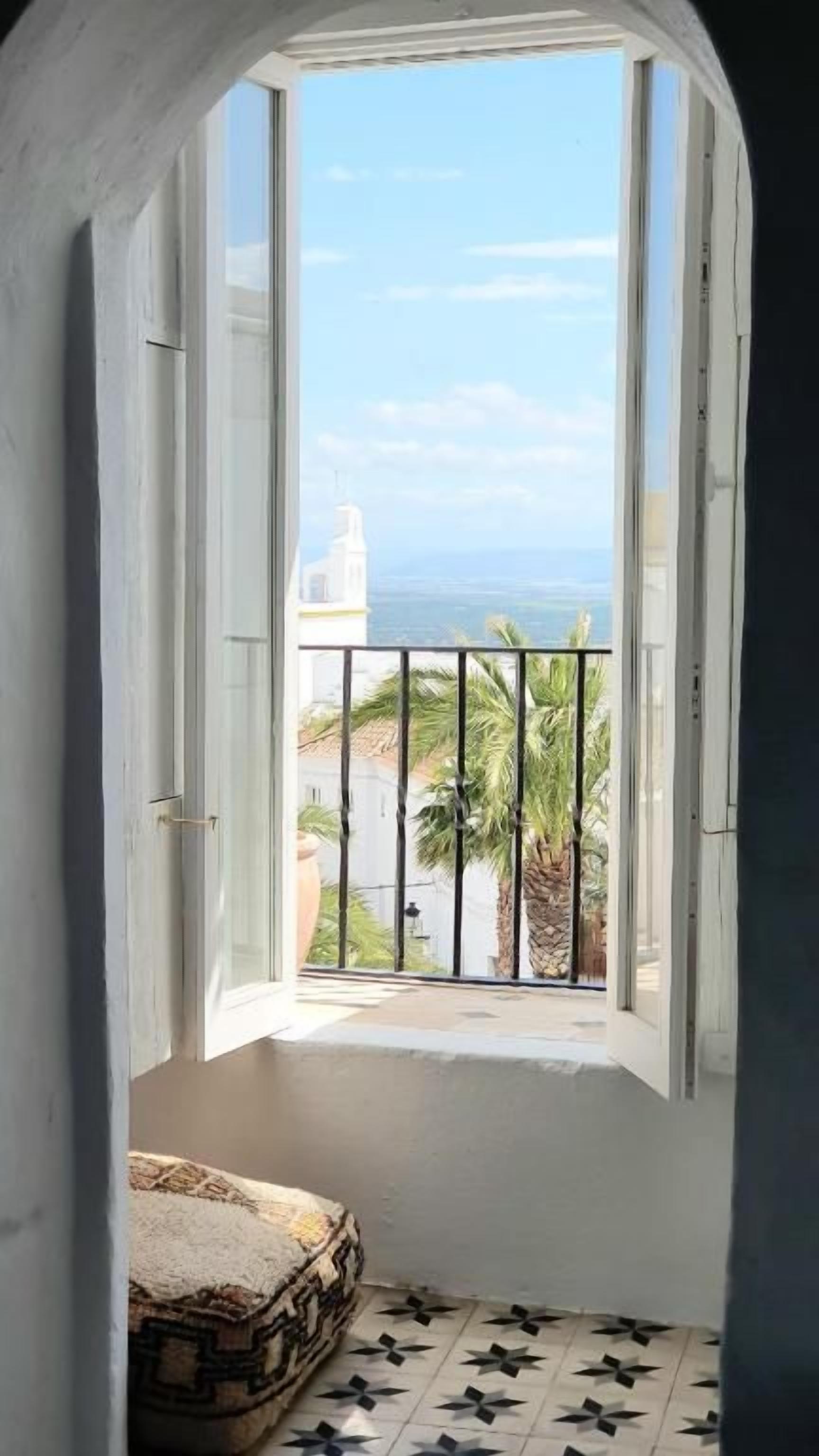 Ein Zimmer mit Meerblick durch ein offenes Fenster, ein Balkon mit Geländer und ein gemusterter Boden.