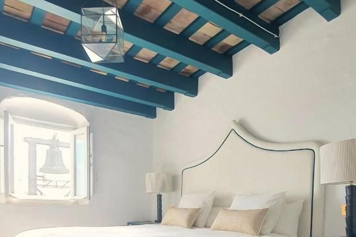 A bedroom with a bed, a nightstand, a lamp, a window, and a wooden ceiling.