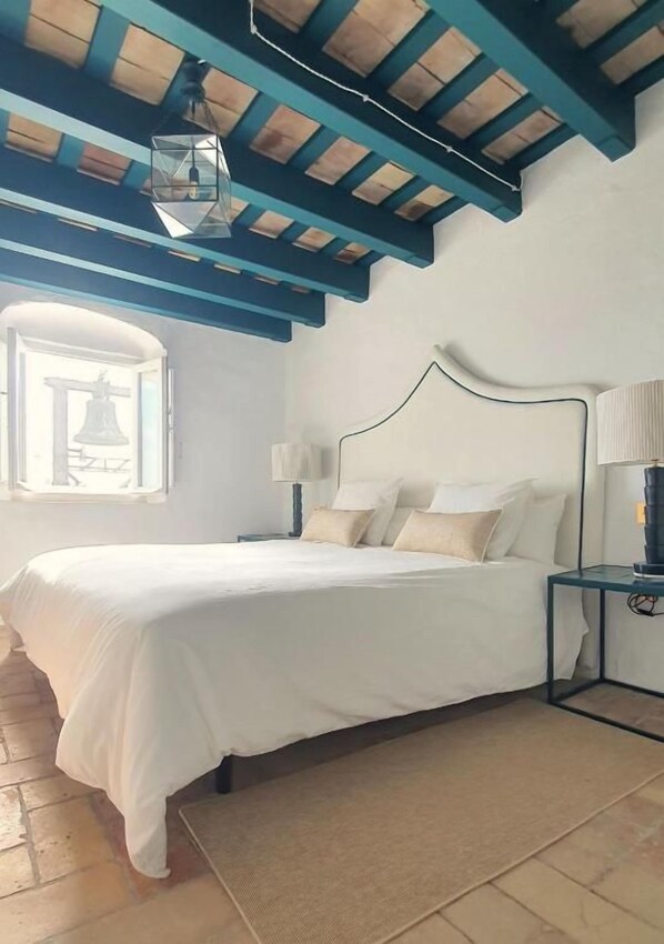 A bedroom with a bed, a nightstand, a lamp, a window, and a wooden ceiling.
