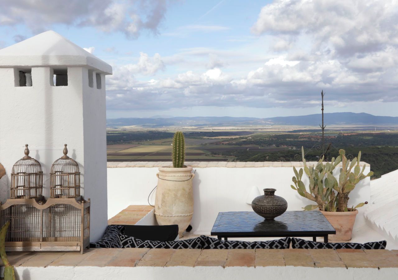 Una terraza con pared blanca, una mesa, dos cactus en macetas, una jaula de pájaros y vistas al campo.