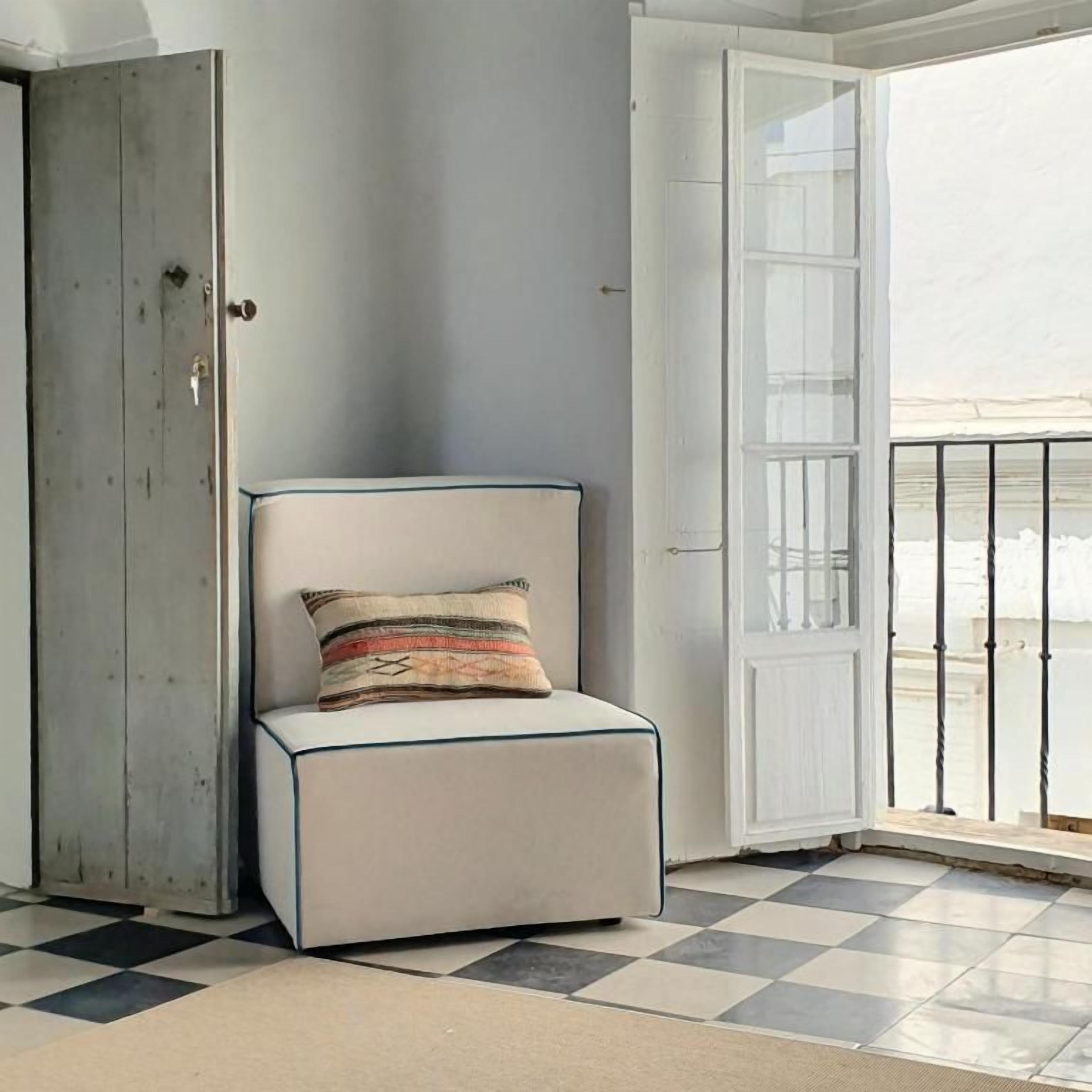 Ein Zimmer mit Schachbrettfußboden, ein weiß gepolsterter Sessel mit gemustertem Kissen, eine Holztür und eine weiße Wand.