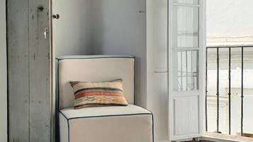 Ein Zimmer mit Schachbrettfußboden, ein weiß gepolsterter Sessel mit gemustertem Kissen, eine Holztür und eine weiße Wand.