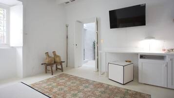 Ein Zimmer mit weißer Wand, einem flachbildfernseher über einem weißen Schrank, einem kleinen weißen Nachttisch und einem gemusterten Teppich auf dem Boden.