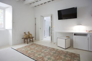 A room with a white wall, a flat-screen TV mounted above a white cabinet, a small white side table, and a patterned rug on the floor.