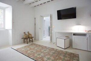 Una habitación con pared blanca, un televisor de pantalla plana montado sobre un mueble blanco, una pequeña mesita auxiliar y una alfombra estampada en el suelo.