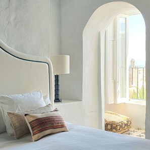A bedroom with a bed, pillows, a lamp, and a view of the outdoors.