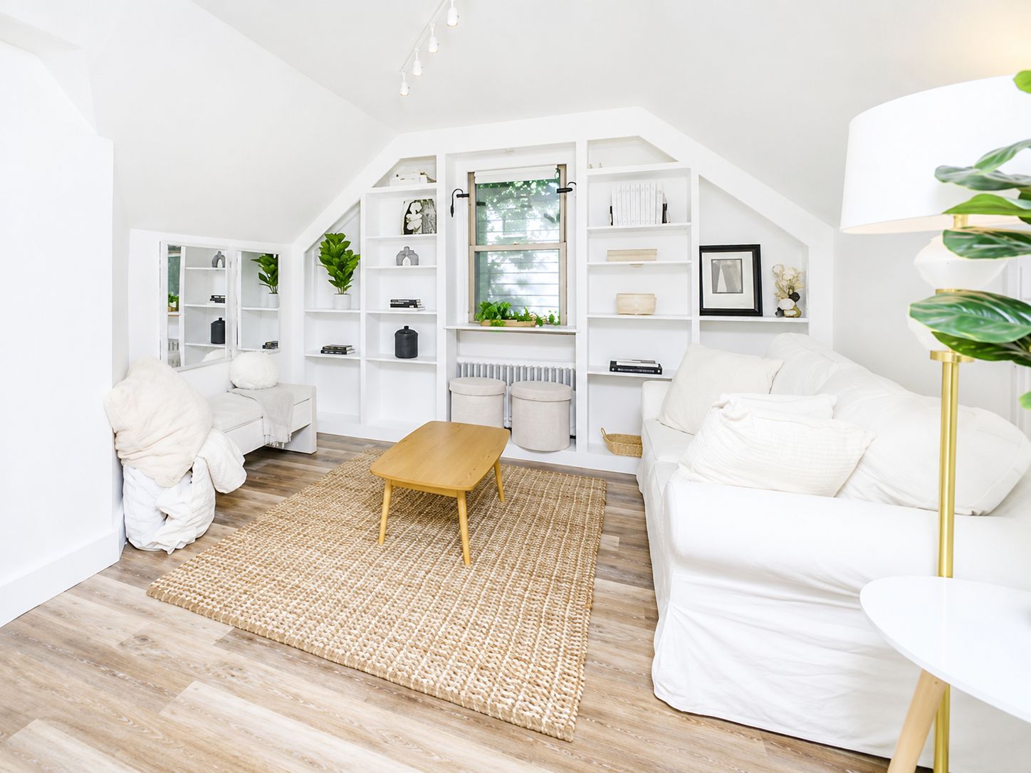 Un salon moderne avec un canapé blanc, une table basse en bois et un fauteuil blanc. On y trouve des étagères blanches où sont disposés divers objets, ainsi qu’une fenêtre donnant sur un paysage verdoyant.