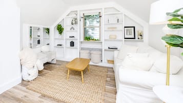 Un salon moderne avec un canapé blanc, une table basse en bois et un fauteuil blanc. On y trouve des étagères blanches où sont disposés divers objets, ainsi qu’une fenêtre donnant sur un paysage verdoyant.