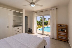 Quarto com cama, ventilador de teto, porta de vidro deslizante para uma piscina e prateleira de madeira.