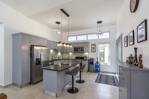 Cozinha moderna com ilha central, banquetas, eletrodomésticos de aço inoxidável, iluminação pendente e vista através de grandes janelas.
