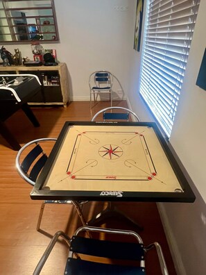 Een eettafel met een spel, stoelen, een raam met jaloezieën en een keukenhoek met een spoelbak en kastjes.