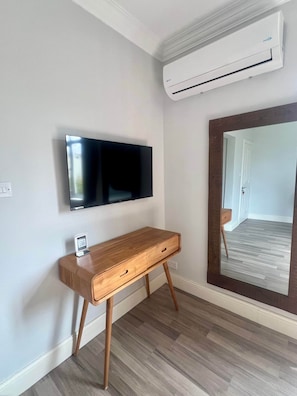 Habitació amb televisió de pantalla plana penjada a la paret, escriptori de fusta amb calaixos i un gran mirall que reflecteix l'espai.