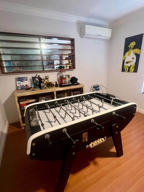 Een kamer met een tafelvoetbaltafel, een houten kast met diverse spullen, een spiegel en een schilderij aan de muur.