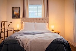 A neatly made bed with a tufted headboard, two bedside tables with lamps, a chair, and a painting on the wall.