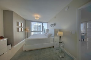 A modern bedroom with a large bed, bedside tables, a window with a city view, and a door leading to another room. - Spectacular 46th Floor Corner 2BR Bay & City View (Miami)