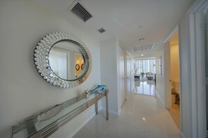 A hallway with a round mirror, a console table, and a view into a room with a toilet and a window. - Spectacular 46th Floor Corner 2BR Bay & City View (Miami)