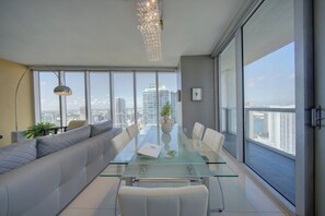 A modern interior with a glass-top table, white leather seating, and large windows offering a city view. - Spectacular 46th Floor Corner 2BR Bay & City View (Miami)