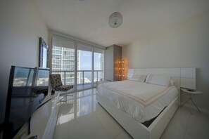 A modern bedroom with a large bed, a TV, a chair, and a view of the city through floor-to-ceiling windows. - Spectacular 46th Floor Corner 2BR Bay & City View (Miami)