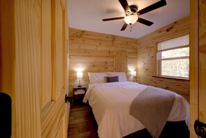 A wooden cabin-style room with a bed, two bedside lamps, a ceiling fan, and a window with blinds. - Ubuntu Haus • Near Castle Rock Lake • HOT TUB! (Friendship)