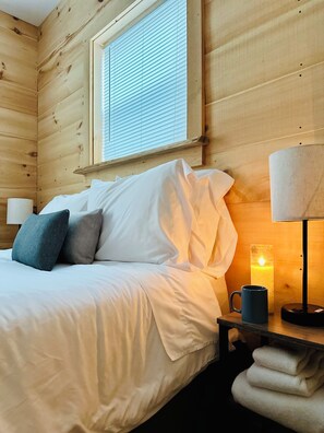 Ein Zimmer aus Holz mit einem Bett, Kissen, einem Nachttisch mit Lampe, einer Kerze und einer Tasse.