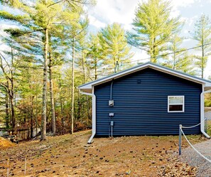 A single-story building with a gabled roof, surrounded by a forest. There are visible electrical boxes and a window on the side of the building. - Ubuntu Haus • Near Castle Rock Lake • HOT TUB! (Friendship)