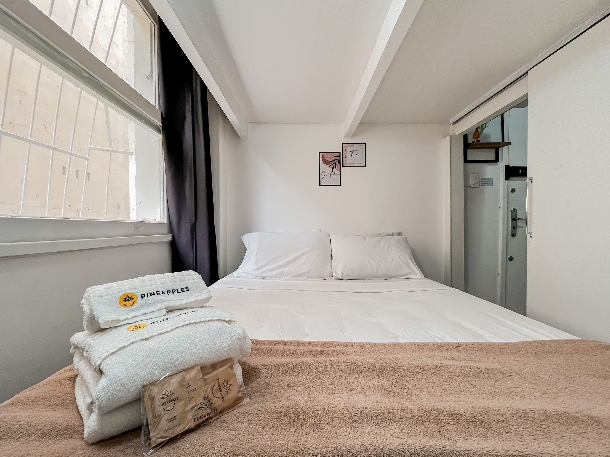 A hotel room with a bed, a window with curtains, a wall with framed artwork, and a door.