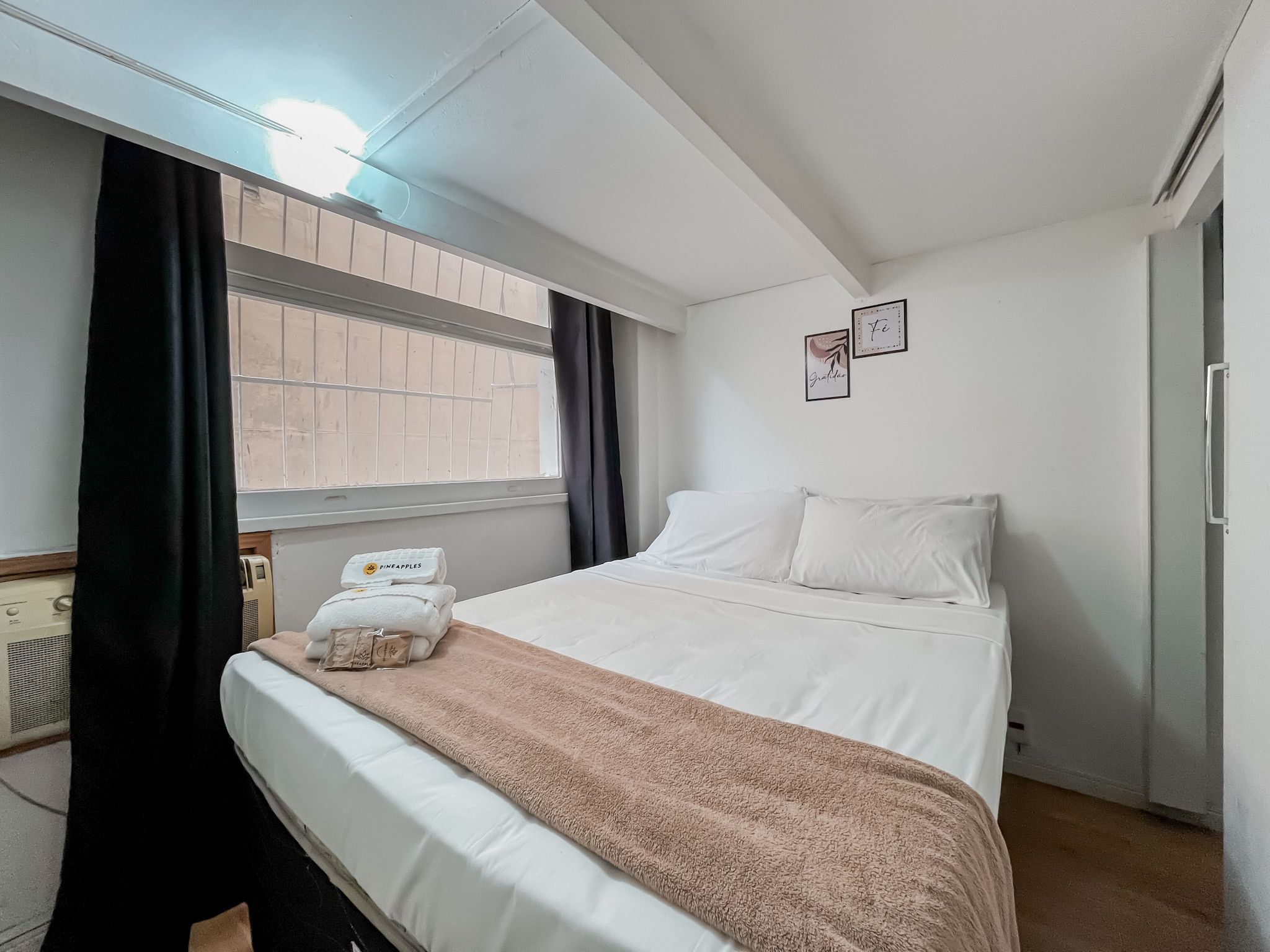 A hotel room with a bed, a window with curtains, a radiator, and a wall with two framed pictures.